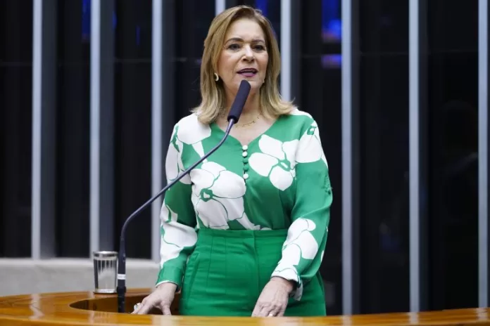 Deputada Lêda Borges fala ao microfone. Ela é branca, loira, usa uma camisa verde com flores brancas