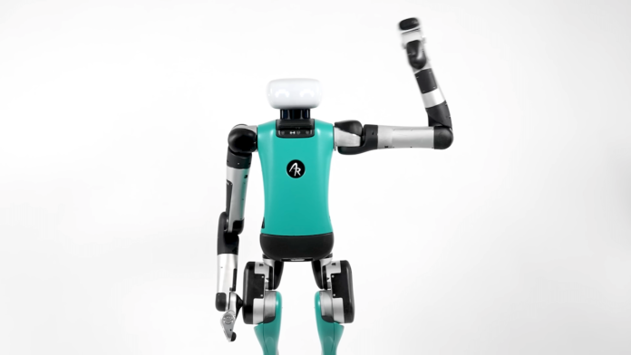 A imagem mostra o robô Digit, da Agility Robotics. Ele é um dispositivo robótico nas cores verde água, cinza e preto, e tem a cabeça branca. Na foto, ele aparece com a mão esquerda levantada, como se fizesse uma saudação.