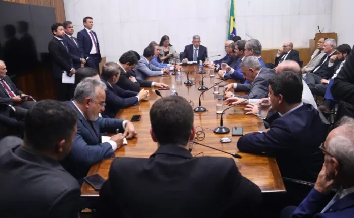 Presidente da Câmara dos Deputados, Arthur Lira, está sentado à mesa conversando com várias pessoas