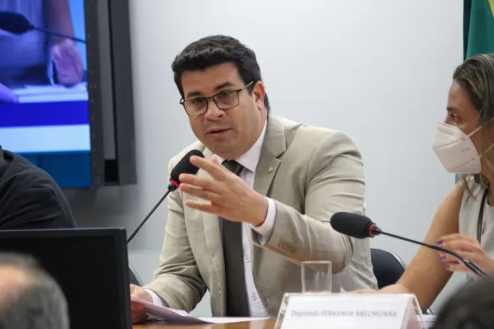 Deputado Carlos Veras fala ao microfone. Ele é branco, tem o cabelo escuro, usa um terno bege e óculos