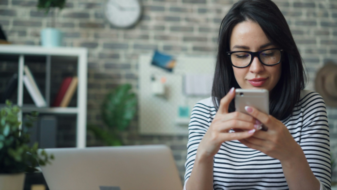 imagem mostra uma mulher sentada em frente a um laptop enquanto mexe em seu celular